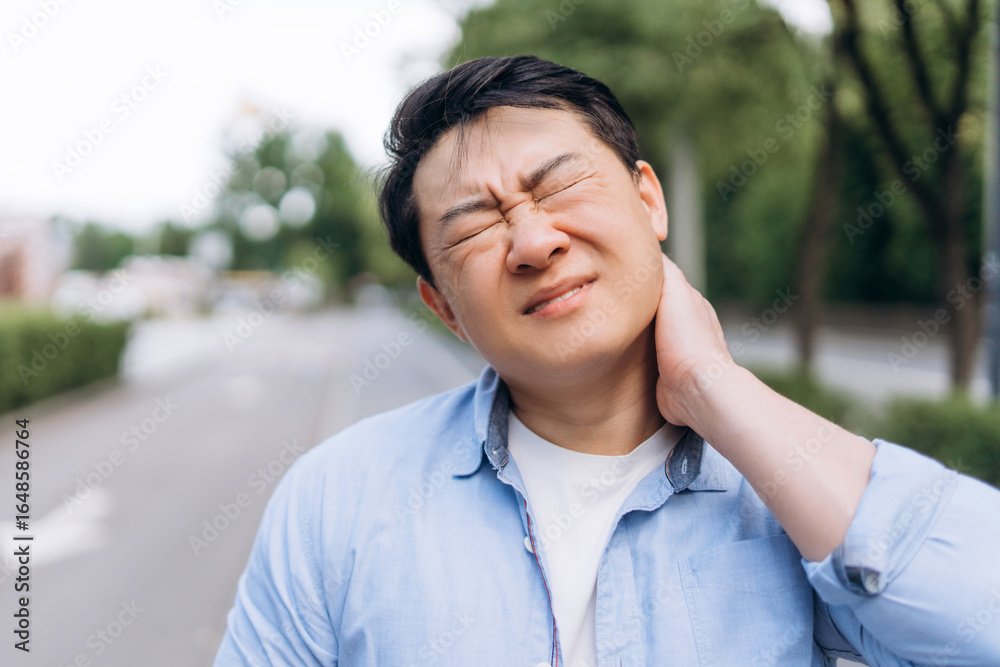 Asian man suffering from neck pain outdoors: grimacing in discomfort