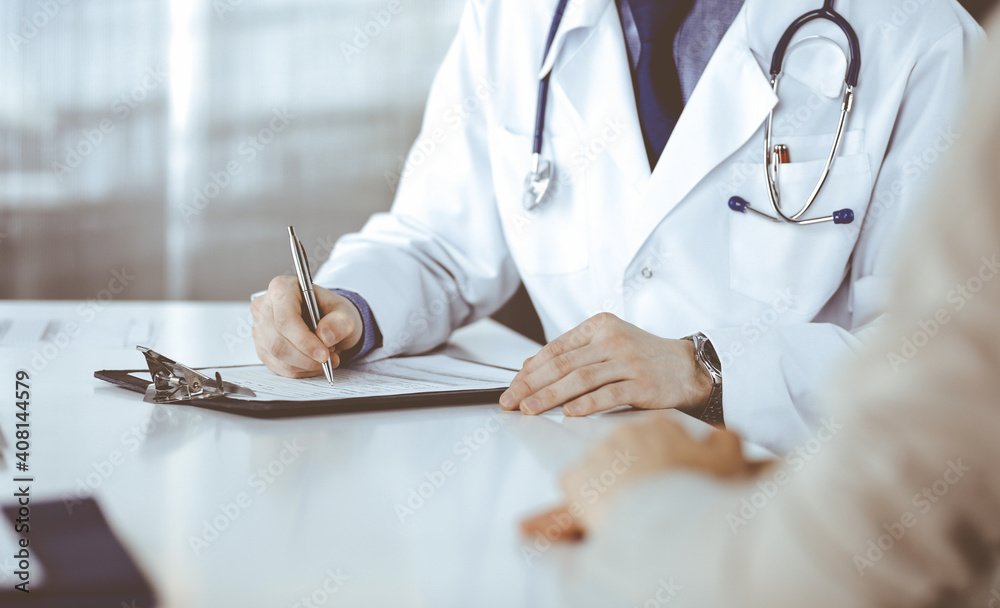 Unknown male doctor and patient woman discussing something while sitting in clinic and using clipboard. Best medical service in hospital, medicine, pandemic stop